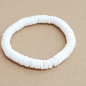 Könnte beinhalten: Ein weißes Armband aus kleinen, runden, scheibenförmigen Perlen. Die Perlen sind zu einer kreisförmigen Form aufgereiht. Das Armband ist vor einem neutralen Hintergrund platziert.