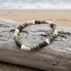 Könnte beinhalten: Ein Perlenarmband mit einem sich wiederholenden Muster aus kleinen, zylindrischen Perlen in den Farben Olivgrün, Dunkelbraun, Hellbeige und leuchtendem Weiß. Die Perlen sind kreisförmig aufgereiht und bilden ein schlichtes, einheitliches Design.