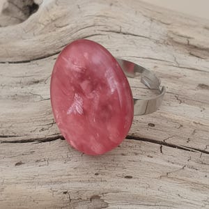 May include: A pink oval gemstone ring with a silver adjustable band. The gemstone has a marbled appearance with varying shades of pink. The ring is set against a weathered wooden background.
