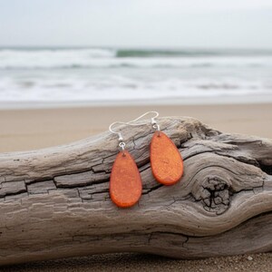 Op de afbeelding: Een paar oranje druppelvormige oorbellen met zilveren haken rust op een verweerd stuk drijfhout. De oorbellen hebben een textuur en zijn geplaatst tegen een achtergrond van een zandstrand en oceaangolven.