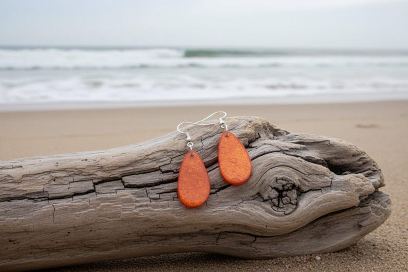 Op de afbeelding: Een paar oranje druppelvormige oorbellen met zilveren haken rust op een verweerd stuk drijfhout. De oorbellen hebben een textuur en zijn geplaatst tegen een achtergrond van een zandstrand en oceaangolven.