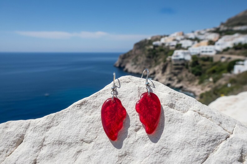 May include: A pair of red, heart-shaped earrings with silver hooks. The earrings are displayed on a white rock, with a scenic background of the ocean and a coastal town under a bright blue sky. Jewelry.