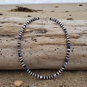 Könnte beinhalten: Eine Halskette mit schwarzen, weißen und blauen Perlen. Die Halskette besteht aus abwechselnd schwarz-weiß gestreiften Perlen, die mit blauen und schwarzen Perlen durchsetzt sind. Die Halskette wird auf einem Stück Treibholz präsentiert.