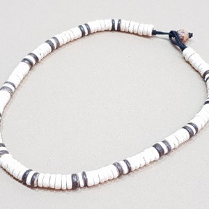 May include: White disc bead necklace with dark gray accent beads. The necklace is strung on a black cord and has a natural bead closure. This necklace is a casual accessory.