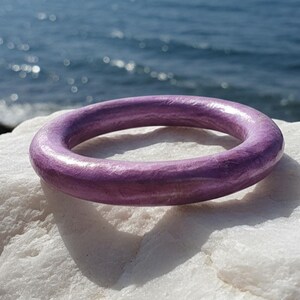 Op de afbeelding: Een ronde, paarse armband ligt op een wit, gestructureerd oppervlak met de oceaan op de achtergrond. De armband is effen van kleur en lijkt gemaakt van hout of een vergelijkbaar materiaal. De armband is een bangle-stijl.