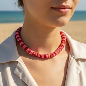Könnte beinhalten: Eine farbenfrohe Halskette aus runden, flachen Perlen in Rosa-, Rot- und Orangetönen. Die Halskette wird um den Hals getragen, mit einem Strand und Meer im Hintergrund. Ein auffälliges Schmuckstück.