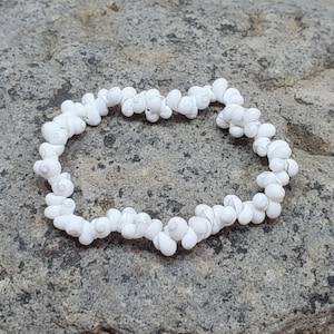 Könnte beinhalten: Ein weißes Muschelarmband. Das Armband besteht aus kleinen, weißen Muscheln, die aneinandergereiht sind. Die Muscheln haben ein Spiralmuster. Das Armband liegt auf einer grauen, strukturierten Oberfläche.