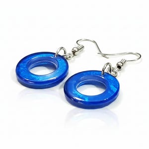 K&ouml;nnte beinhalten: Ein Paar blaue Resin-Creolen mit silberfarbenen Ohrhaken. Die Ohrringe haben eine gl&auml;nzende Oberfl&auml;che und ein kreisf&ouml;rmiges Design. Das Blau hat einen schimmernden Effekt, der dem Schmuck visuelles Interesse verleiht.