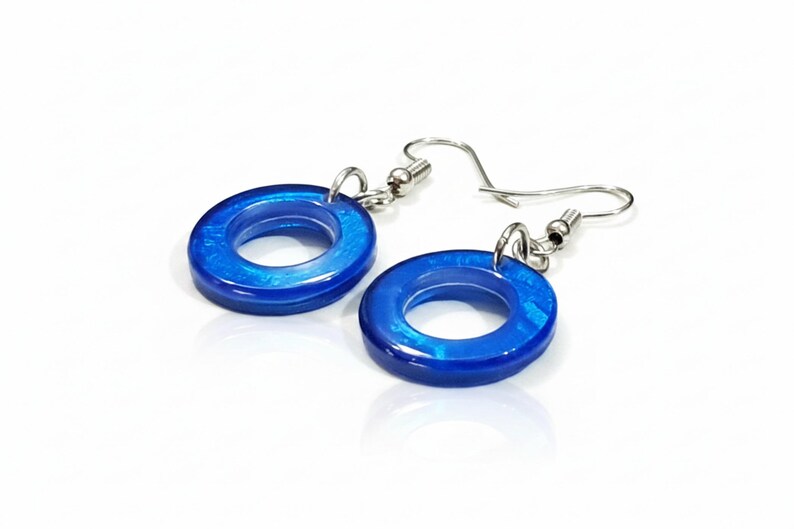 K&ouml;nnte beinhalten: Ein Paar blaue Resin-Creolen mit silberfarbenen Ohrhaken. Die Ohrringe haben eine gl&auml;nzende Oberfl&auml;che und ein kreisf&ouml;rmiges Design. Das Blau hat einen schimmernden Effekt, der dem Schmuck visuelles Interesse verleiht.