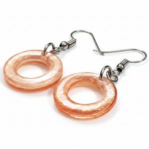 K&ouml;nnte beinhalten: Ein Paar Ohrringe mit einem kreisf&ouml;rmigen, donut-f&ouml;rmigen Design in transluzenter Pfirsichfarbe. Die Ohrringe haben silberfarbene Haken und Verbindungsteile. Der Schmuck ist vor einem wei&szlig;en Hintergrund platziert.