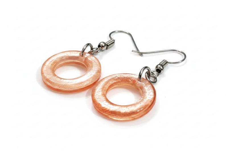 K&ouml;nnte beinhalten: Ein Paar Ohrringe mit einem kreisf&ouml;rmigen, donut-f&ouml;rmigen Design in transluzenter Pfirsichfarbe. Die Ohrringe haben silberfarbene Haken und Verbindungsteile. Der Schmuck ist vor einem wei&szlig;en Hintergrund platziert.