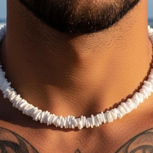 Puede incluir: Collar de conchas blancas. El collar está hecho de pequeñas conchas blancas de forma irregular ensartadas. El collar se lleva alrededor del cuello. El fondo muestra una vista borrosa del océano.