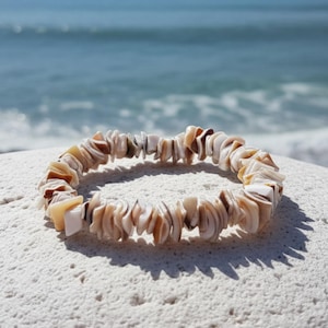 Op de afbeelding: Een armband van natuurlijke schelpen met een crème, bruine en beige kleurenschema. De armband is gemaakt van kleine, onregelmatig gevormde schelpfragmenten. De achtergrond toont een wit oppervlak en de zee.