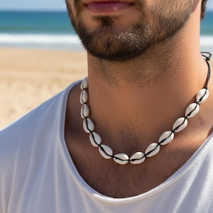 Könnte beinhalten: Halskette aus weißen Muscheln mit schwarzem Faden. Die ovalen Muscheln sind mit einem schwarzen Faden verbunden. Die Person trägt ein weißes T-Shirt. Im Hintergrund sind Strand und Meer zu sehen.