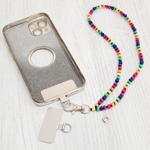Könnte beinhalten: Eine transparente Handyhülle mit Glitzer und einer silbernen Metallplatte sowie einem farbenfrohen Perlenband. Das Band weist eine Vielzahl von Farben auf, darunter Rot, Orange, Gelb, Grün, Blau und Lila.
