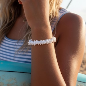 Peut inclure: Bracelet en pierres blanches. Le bracelet est composé de petites pierres blanches de forme irrégulière, enfilées ensemble. Il est porté sur un bras bronzé, avec un haut rayé bleu et blanc en arrière-plan.