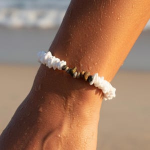 Op de afbeelding: Een kralenarmband bestaande uit een reeks kleine, witte, onregelmatig gevormde schelpachtige kralen, afgewisseld met een centrale groep gladde, aardekleurige stenen in tinten bruin, grijs en zwart, gerangschikt in een cirkelvormige band.