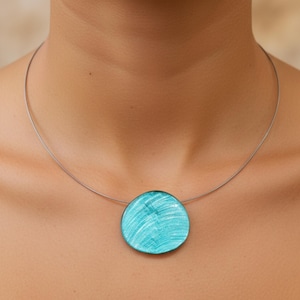 Op de afbeelding: Een minimalistische ketting met een dunne, zilverkleurige draad en een ronde, turquoise hanger. De hanger heeft een textuur met witte penseelstreken. Een eenvoudige maar elegante accessoire.