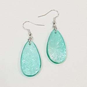 Op de afbeelding: Een paar druppelvormige oorbellen. Elke oorbel heeft een doorschijnende, lichtblauwe hanger met een textuur. Ze hangen aan zilverkleurige oorhaakjes. De oorbellen worden getoond tegen een witte achtergrond.