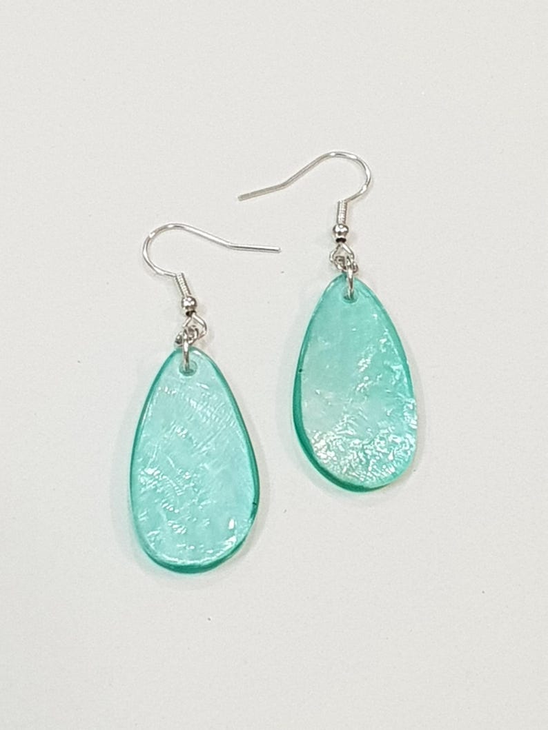 Op de afbeelding: Een paar druppelvormige oorbellen. Elke oorbel heeft een doorschijnende, lichtblauwe hanger met een textuur. Ze hangen aan zilverkleurige oorhaakjes. De oorbellen worden getoond tegen een witte achtergrond.