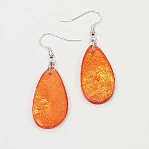 Op de afbeelding: Een paar oranje druppelvormige oorbellen met zilverkleurige oorhaken. De oorbellen hebben een getextureerde, iriserende afwerking en worden gepresenteerd tegen een witte achtergrond. Deze oorbellen zijn een stijlvol accessoire.