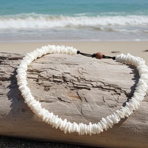 Pode incluir: Um colar de conchas brancas com um cordão preto e fecho de contas de madeira repousa sobre um pedaço de madeira flutuante. O colar é composto por pequenos pedaços irregulares de concha, criando uma estética texturizada e praiana. O oceano e a praia são visíveis ao fundo.