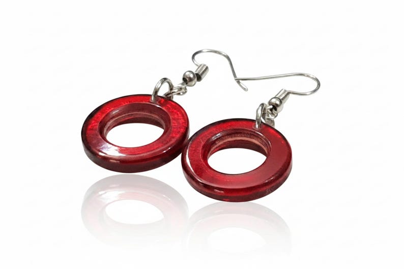 K&ouml;nnte beinhalten: Ein Paar rote, kreisf&ouml;rmige Ohrringe mit silberfarbenen Haken. Die Ohrringe haben eine gl&auml;nzende Oberfl&auml;che und spiegeln sich auf einer wei&szlig;en Oberfl&auml;che. Ein schlichtes, aber elegantes Accessoire.