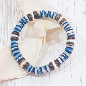 May include: A beaded bracelet featuring a circular design. The bracelet is composed of small, flat, round beads in shades of blue, brown, and white. The bracelet is resting on a white, textured surface.