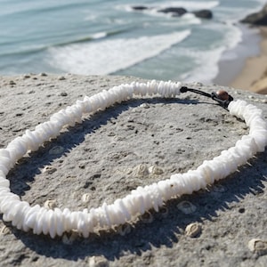 May include: A white shell necklace lies on a grey rock, with the sea in the background. The necklace is made of small, irregular white shells strung together. The necklace is approximately 40 cm long.