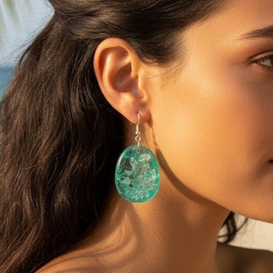 Puede incluir: Primer plano de un pendiente de plata con un colgante grande, ovalado, de color turquesa. El colgante tiene un diseño veteado blanco y turquesa. El fondo está borroso, con toques de azul y verde.