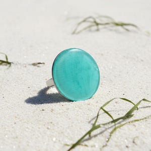 May include: A round, turquoise ring with a silver band rests on white sand. The ring's large, circular turquoise stone is the focal point, with a smooth, glossy surface. Green sea grass is scattered around the ring.