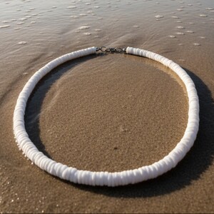 Könnte beinhalten: Eine weiße Halskette aus kleinen, scheibenförmigen Perlen. Die Halskette liegt an einem Sandstrand, wobei das Meerwasser sie sanft berührt. Die Halskette hat einen silberfarbenen Verschluss.