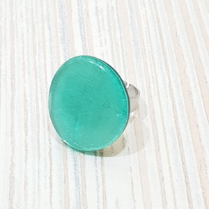 Könnte beinhalten: Verstellbarer Ring mit silberfarbenem Band und einem großen, runden, türkisfarbenen Kunstharzstein. Der Stein hat eine glatte, glänzende Oberfläche. Der Ring wird auf einer hellen, vertikal gestreiften Oberfläche gezeigt.
