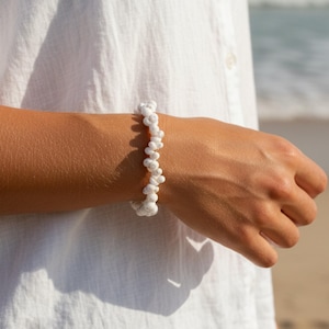 Könnte beinhalten: Ein Armband bestehend aus zahlreichen kleinen, weißen, spiralförmigen Muscheln, die in einer dichten, durchgehenden Reihe aufgereiht sind. Die Muscheln sind einheitlich in Farbe und Textur und bilden ein strukturiertes, zylindrisches Band.