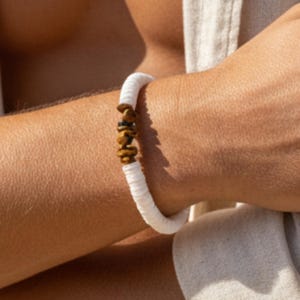 Könnte beinhalten: Ein Armband aus kleinen, flachen, weißen, kreisförmigen Perlen, die in einem durchgehenden Band angeordnet sind, mit einem Mittelteil aus drei unregelmäßigen, texturierten, goldbraunen Steinperlen, die einen deutlichen Kontrast bilden.