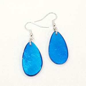 Op de afbeelding: Een paar helderblauwe druppelvormige oorbellen met zilverkleurige haken. De oorbellen hebben een glanzende afwerking en reflecteren het licht. Ze worden gepresenteerd tegen een witte achtergrond.