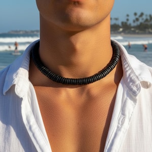 Könnte beinhalten: Eine schwarze Perlenkette aus kleinen, runden Perlen wird um den Hals getragen. Die Kette ist vor einem weißen Hemd zu sehen. Der Hintergrund zeigt eine Strandszene mit Meer und Menschen.