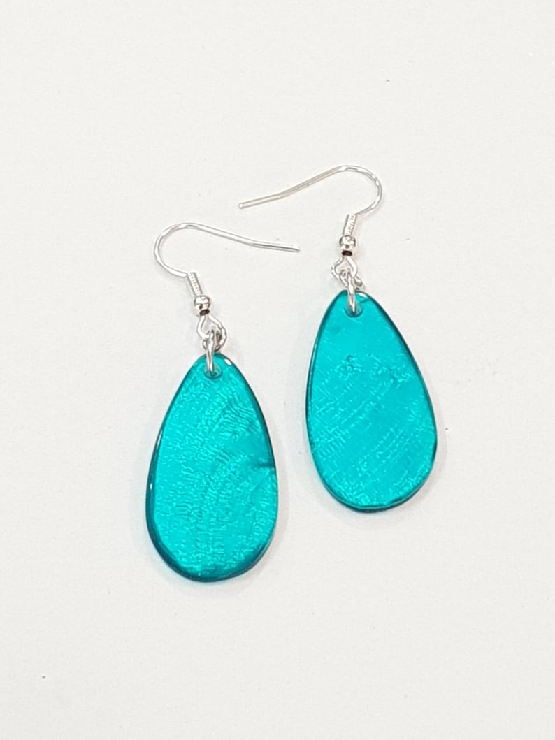 Op de afbeelding: Een paar turquoise teardrop oorbellen met zilverkleurige oorhaken. De oorbellen hebben een getextureerde, iriserende afwerking die licht reflecteert. De oorbellen worden gepresenteerd tegen een witte achtergrond.