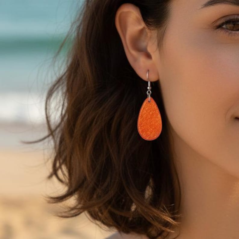 Op de afbeelding: Close-up van een oor van een vrouw met een enkele oranje druppelvormige oorbel. De oorbel is een effen oranje kleur met textuur en hangt aan een zilveren haak. De achtergrond is wazig.