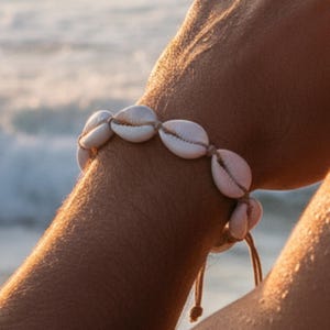 Peut inclure: Un bracelet composé d'une série de coquillages cauris blancs enfilés sur un cordon beige. Les coquillages sont disposés en rangée continue, avec leurs ouvertures naturelles orientées vers l'extérieur. Le cordon est noué avec des extrémités.