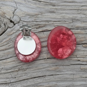 Könnte beinhalten: Ein Paar Clip-Ohrringe mit rotem, marmoriertem Design. Ein Ohrring hat einen silbernen Clip auf der Rückseite. Die Ohrringe sind oval und haben ein glänzendes Finish. Sie liegen auf einer verwitterten, grauen Holzoberfläche.