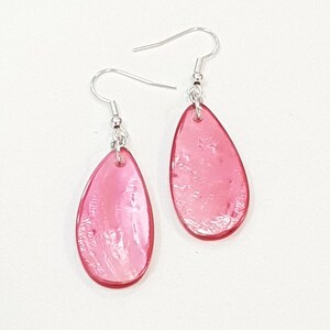 Op de afbeelding: Een paar roze druppelvormige oorbellen met zilverkleurige haken. De oorbellen hebben een doorschijnende, parelmoerachtige uitstraling en de druppelvorm is omlijnd met een donkerdere roze tint. Witte achtergrond.