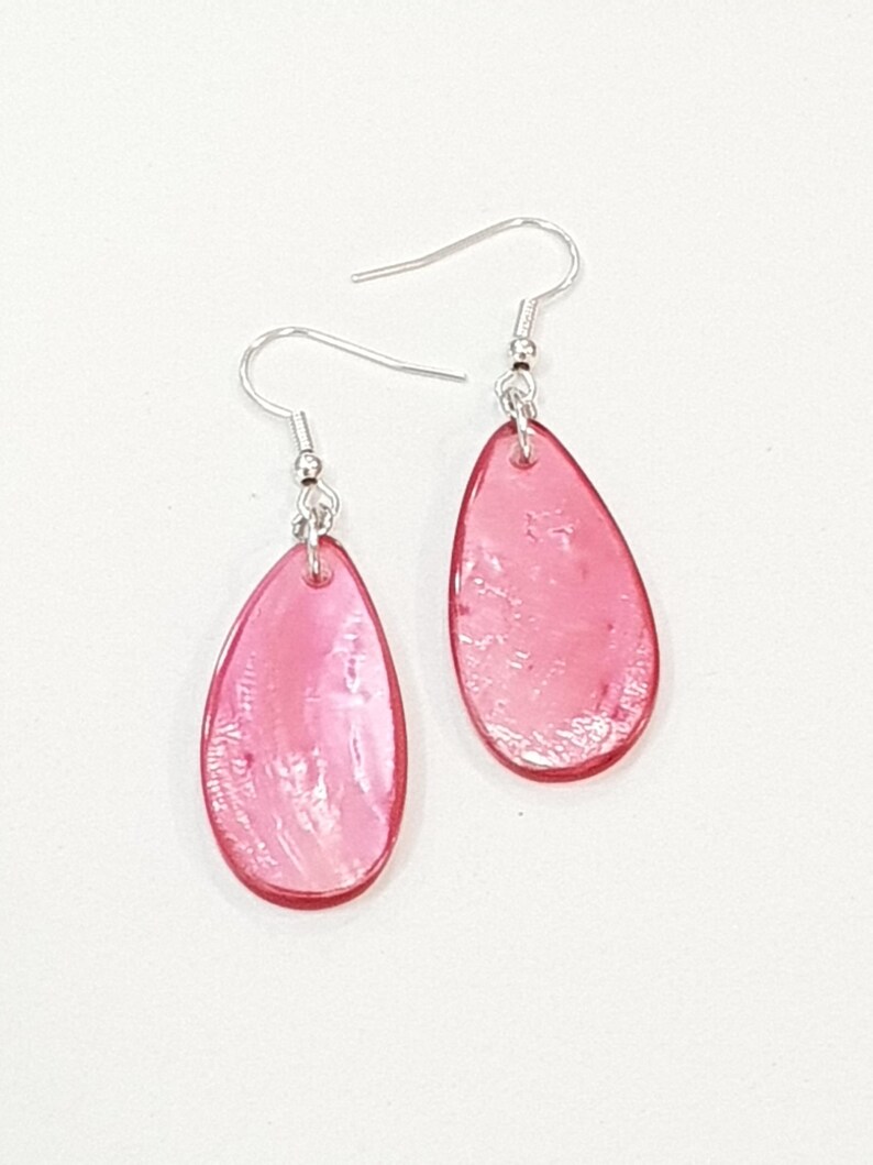 Op de afbeelding: Een paar roze druppelvormige oorbellen met zilverkleurige haken. De oorbellen hebben een doorschijnende, parelmoerachtige uitstraling en de druppelvorm is omlijnd met een donkerdere roze tint. Witte achtergrond.