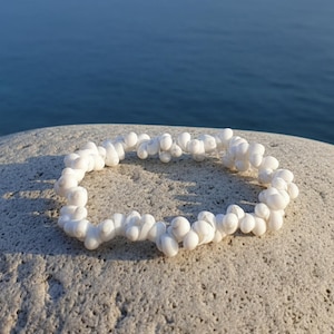 Könnte beinhalten: Ein weißes Perlenarmband aus kleinen, abgerundeten Perlen. Das Armband liegt auf einem strukturierten, hellfarbigen Stein, mit einem verschwommenen blauen Meer im Hintergrund. Die Perlen scheinen aus einem glänzenden Material zu bestehen.