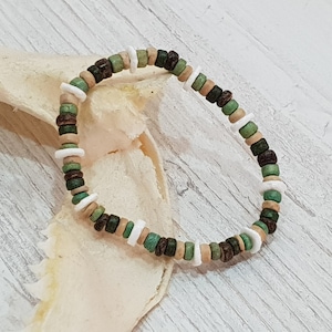 Könnte beinhalten: Ein Armband mit Perlen aus einer Mischung aus Naturholz, grünen, weißen und dunkelbraunen Perlen. Das Armband besteht aus kleinen, runden Perlen, die zusammen aufgefädelt sind und ein lässiges, erdiges Accessoire bilden. Das Armband wird auf einer hellen Oberfläche gezeigt.