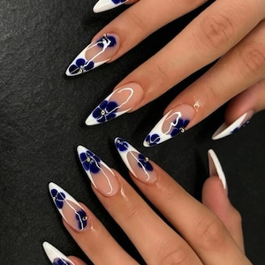 Può includere: Unghie lunghe a forma di mandorla con stile french manicure, punte bianche e base trasparente. Ogni unghia è decorata con motivi floreali blu scuro e piccoli borchie metalliche dorate posizionate al centro dei fiori come dettaglio.