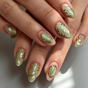Puede incluir: Uñas de forma ovalada con una base de color verde salvia, detalles de hojas de oro metálico y pequeños motivos florales de color rosa claro. Algunas uñas incluyen un marco dorado texturizado en relieve alrededor de un centro verde salvia.