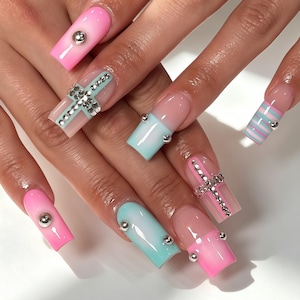 May include: Long square-shaped artificial nails featuring a mix of pastel pink and light blue ombre gradients, horizontal stripes, and clear sections. The design is accented with silver metallic studs and cross-shaped rhinestone embellishments.