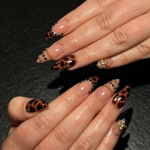 Puede incluir: Uñas puntiagudas en forma de almendra con una base de esmalte nude y patrones de estampado de jirafa en marrón. El diseño incluye detalles de cadenas doradas, pequeños diamantes de imitación negros en las cutículas y dijes de mariposa dorados.