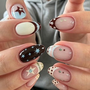 Può includere: Unghie di forma ovale con un mix di design, tra cui una base marrone scuro con stelle azzurre, una base azzurra con una stella marrone scuro, punte bianche con pois marroni e una base nude rosata con piccoli accenti a forma di stella e punto.